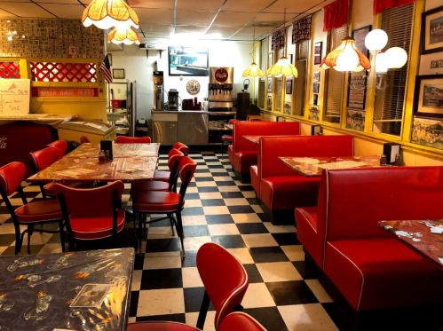 A retro diner interior with red booths, checkered floor, and vintage decor, featuring tables and a TV in the background.
