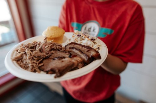 A person holds a plate with sliced brisket, pulled pork, coleslaw, and a cornbread muffin.