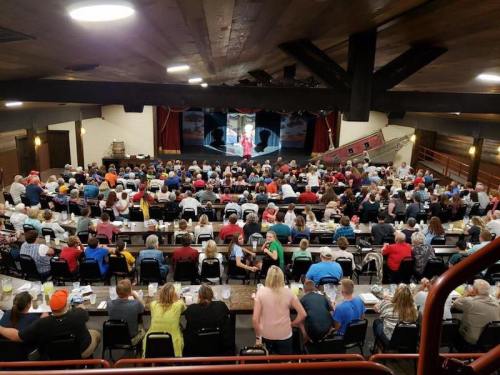 A large audience seated at tables in a theater, watching a performance on stage.