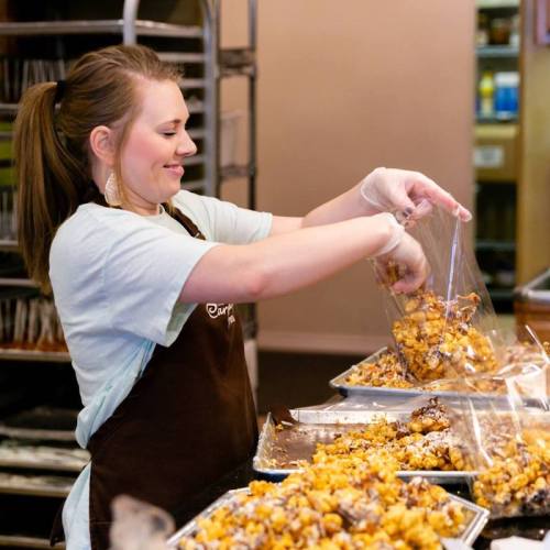 A smiling woman in an apron bags popcorn in a kitchen, surrounded by trays of popcorn treats.