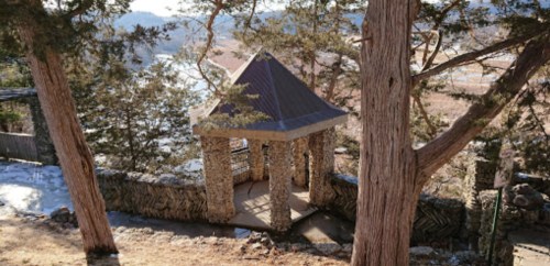 A stone gazebo surrounded by trees, set in a scenic outdoor landscape with snow on the ground.