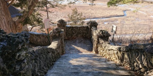 Stone steps lead down through a natural landscape, surrounded by trees and a grassy field in the distance.