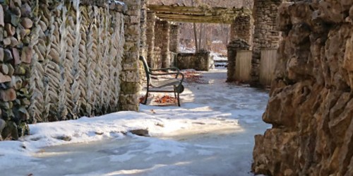A snowy pathway leads through stone ruins, with a bench under a wooden trellis in a serene, winter landscape.