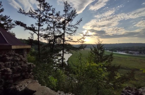 Scenic view of a river and fields at sunset, framed by trees and a stone structure in the foreground.