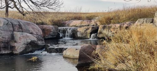A serene landscape featuring a small waterfall flowing over rocks, surrounded by tall grass and a clear sky.