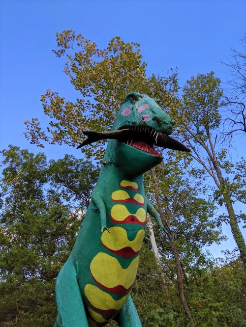 A colorful dinosaur statue with a mustache, surrounded by trees and a clear blue sky.