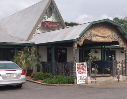 Exterior of a restaurant with a decorative sign, surrounded by greenery and a parking area.