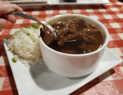 A bowl of beef stew with a spoon, served alongside a portion of white rice on a checkered tablecloth.