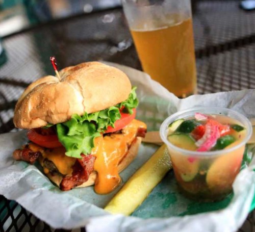 A cheeseburger with lettuce, tomato, and bacon, served with a side of pickles and a cup of mixed vegetables, plus a drink.
