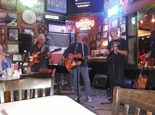 A band performs on stage in a lively bar, with musicians playing guitars and singing under colorful lights.