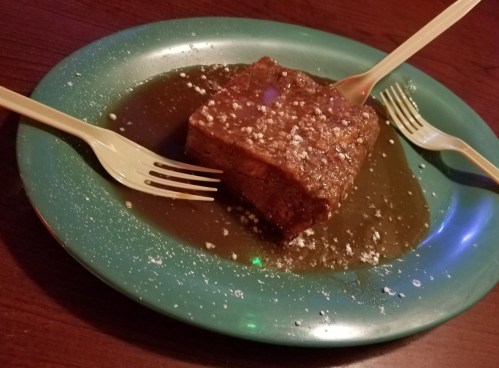A square dessert on a green plate, drizzled with sauce and sprinkled with powdered sugar, with two plastic forks.