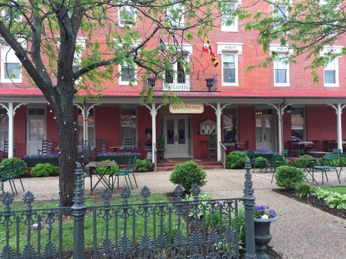 A red-brick building with a porch, surrounded by greenery and outdoor seating, featuring Maryland state flags.