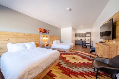 A cozy hotel room featuring two beds, a kitchenette, and modern decor with wooden accents and a colorful rug.