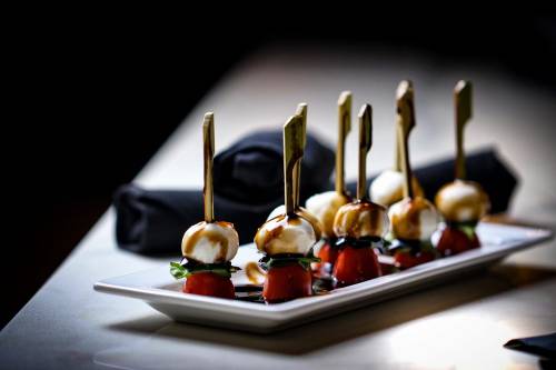A platter of skewered cherry tomatoes and mozzarella balls drizzled with balsamic glaze, garnished with basil.