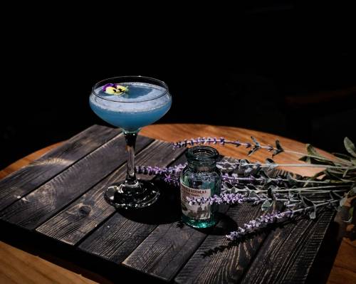 A blue cocktail garnished with a flower, beside a small bottle and lavender sprigs, on a wooden table.