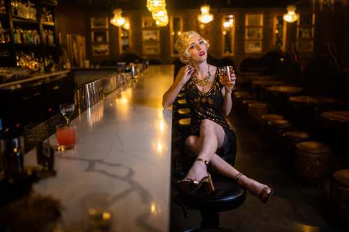 A stylish woman in a black dress sits at a bar, holding a drink, with a glamorous atmosphere and dim lighting.