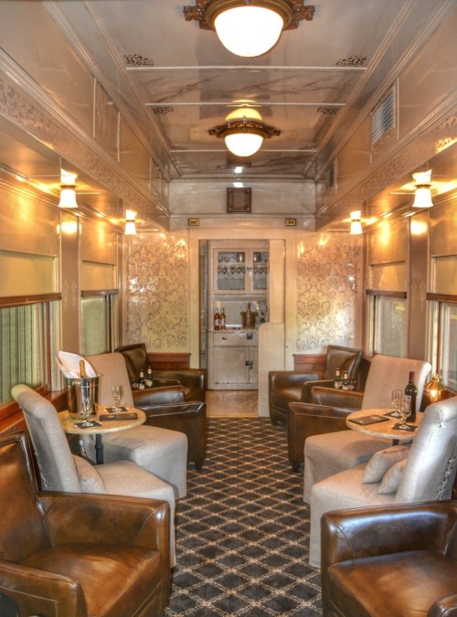 Luxurious train interior featuring plush seating, elegant decor, and a small bar area, illuminated by vintage lighting.