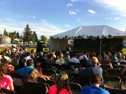 A crowd gathers under a tent for an outdoor event, with a speaker addressing the audience in a park setting.