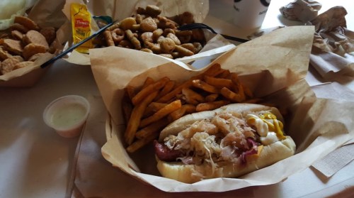 A hot dog topped with sauerkraut and mustard, served with fries and a side of dipping sauce, on a paper tray.