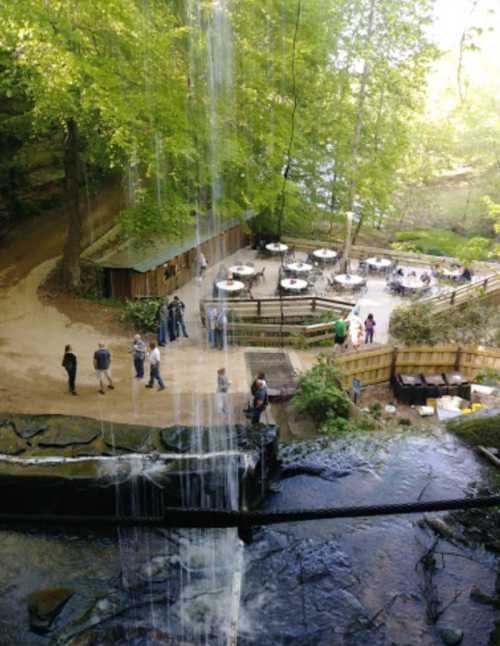 A scenic view of a waterfall cascading over rocks, with people gathered at a nearby outdoor dining area surrounded by trees.