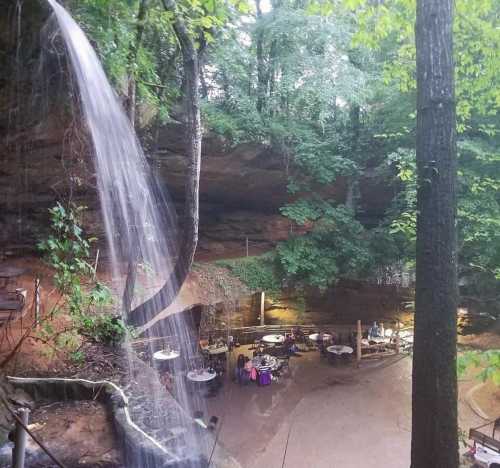 A serene waterfall cascades into a lush, green forest, with a dining area nestled among the trees below.