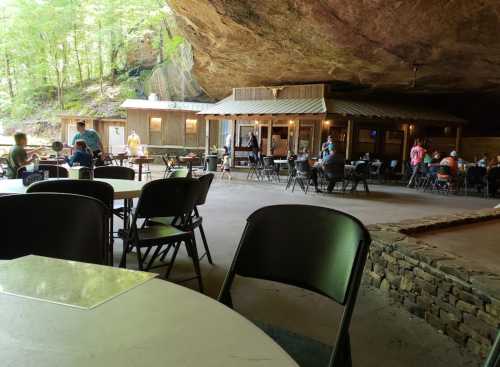 A rustic dining area under a rock overhang, with tables and chairs, and people enjoying their meals in a natural setting.