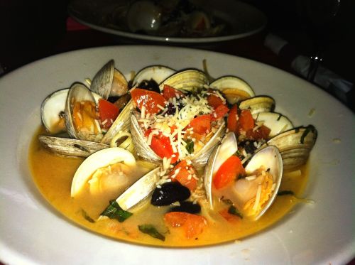 A plate of clams in a creamy broth, garnished with tomatoes, herbs, and cheese, served in a restaurant setting.