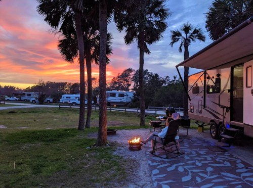 A serene campsite at sunset, featuring a camper, palm trees, and a person relaxing by a fire pit.