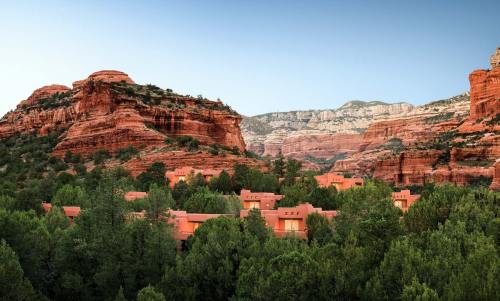 A scenic view of red rock formations and green trees, with adobe-style buildings nestled in the valley.