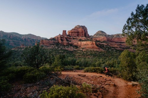 A mountain biker rides along a dirt trail with red rock formations and lush greenery in the background.
