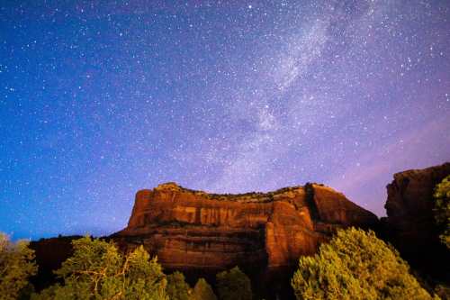Starry night sky over a rugged rock formation, with trees in the foreground and a hint of the Milky Way visible.