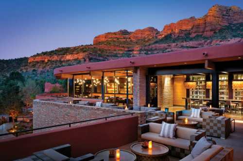 A stylish outdoor lounge area with cozy seating, overlooking red rock formations at sunset. Warm lights create a welcoming ambiance.