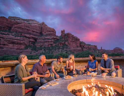 A group of six people enjoying drinks and snacks around a fire pit at sunset, with red rock formations in the background.