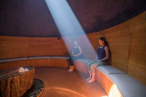Two women sit in a wooden sauna, illuminated by a beam of light, creating a serene and tranquil atmosphere.