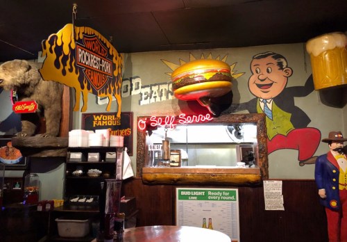 A quirky restaurant interior featuring a large burger sign, a bear, and colorful wall art with a neon "Self Serve" sign.