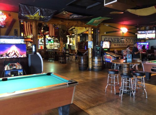 A lively bar interior featuring pool tables, arcade games, and rustic decor with flags and barrels.