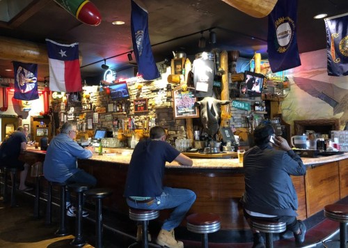 A lively bar scene with patrons sitting at the counter, surrounded by flags and rustic decor.