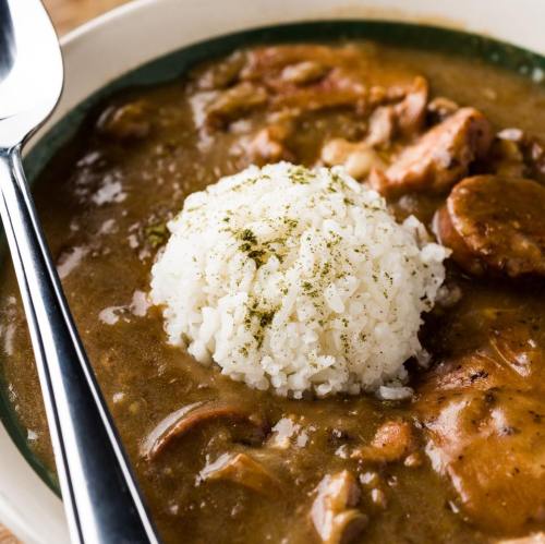 A bowl of savory stew with tender meat and a mound of white rice, garnished with herbs. A spoon rests beside it.