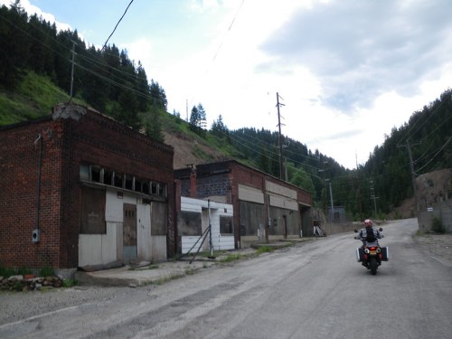Abandoned brick buildings line a rural road, with a motorcycle parked nearby and mountains in the background.