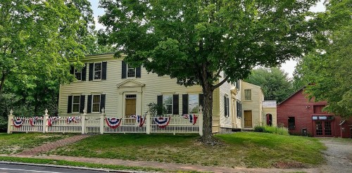 A yellow historic house with a white fence, decorated with red, white, and blue bunting, surrounded by trees.