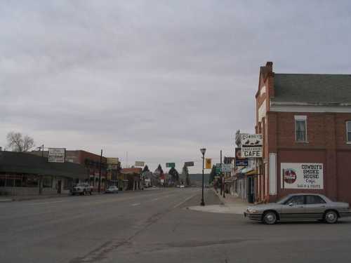 A quiet small-town street lined with shops and a parked car under a cloudy sky.