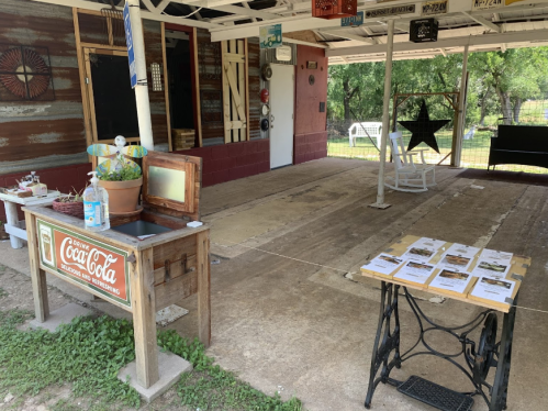 A rustic outdoor space with a Coca-Cola table, a sewing machine, and informational papers on a table under a covered area.