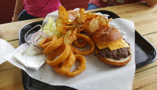 A tray with a cheeseburger, onion rings, potato chips, lettuce, and pickles on a wooden table.