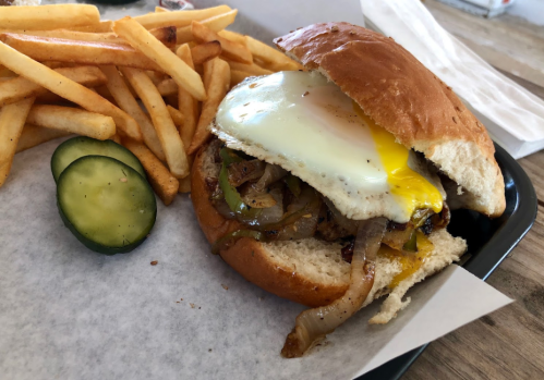 A burger with a fried egg, sautéed onions, and peppers, served with fries and a pickle on the side.