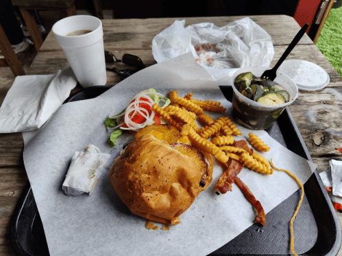 A tray with a burger, crinkle fries, salad, pickles, and a drink, set on a wooden table.