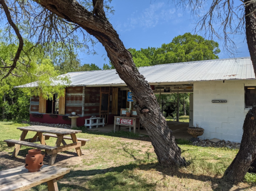 A rustic building with a metal roof, surrounded by trees and picnic tables, set in a grassy area under a blue sky.