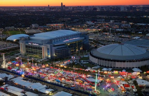 Aerial view of a stadium and fairgrounds illuminated at dusk, with rides and attractions under a colorful sunset.