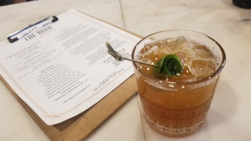 A cocktail with a garnish on a table next to a menu, featuring ice and a light amber color.