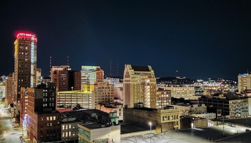 Nighttime cityscape featuring illuminated buildings and a skyline with a clear sky in the background.