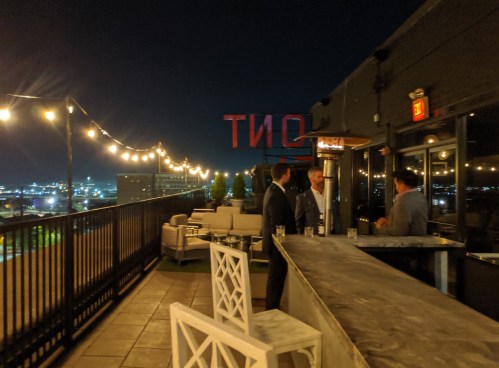 A rooftop bar at night with string lights, a city skyline, and three men chatting near a marble bar.